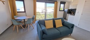 A modern living room with a blue sofa, two yellow cushions, a dining table with four chairs, and a kitchen area with a refrigerator and stove. - NEW T2 NEAR THE PORT AND ALL SHOPS (Porto-Vecchio)