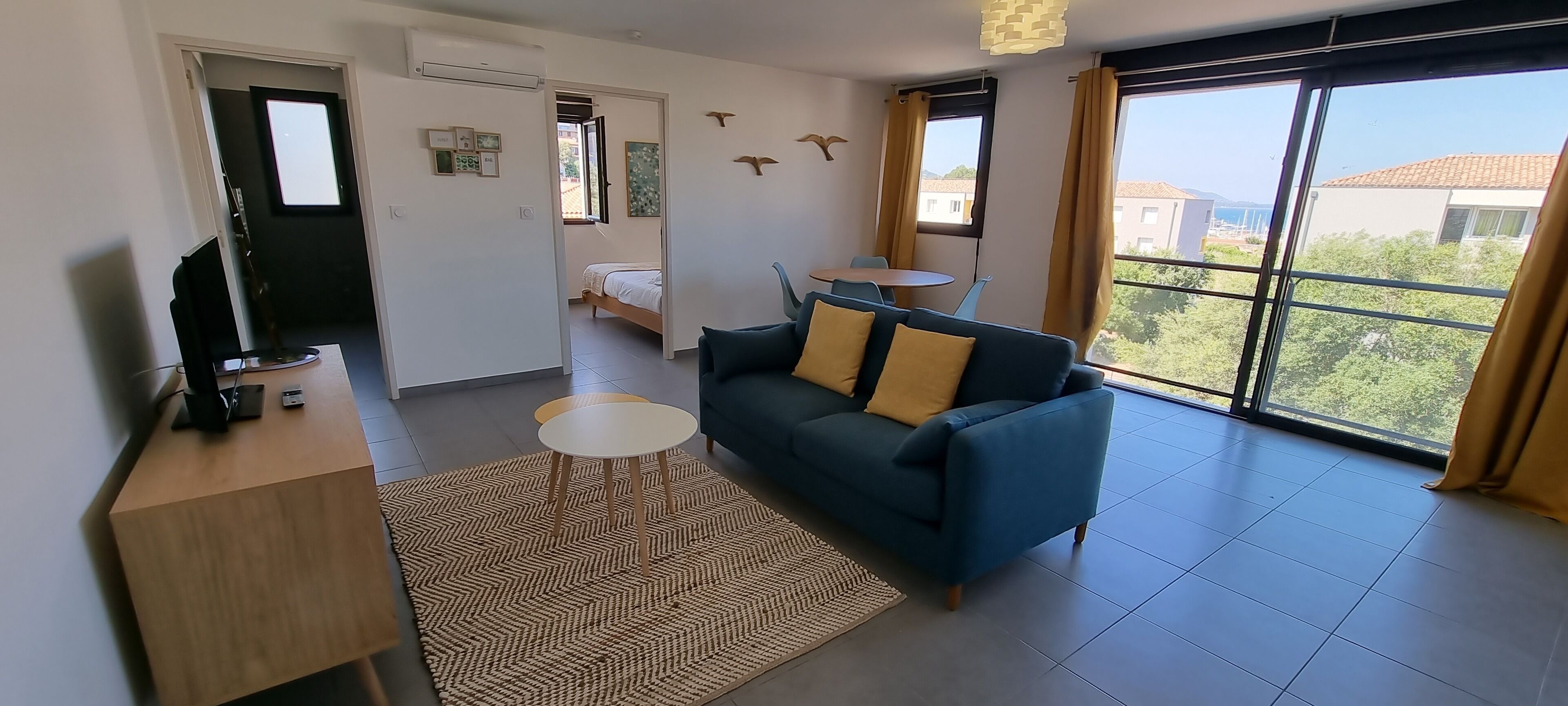 Un salon moderne avec un canapé, une petite table et un téléviseur. Une porte donne accès à une chambre à coucher qui contient un lit et une fenêtre.