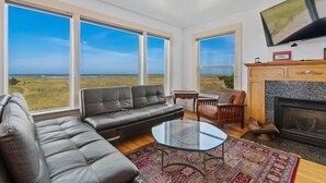 A spacious living room with a large window offering a view of the ocean, a fireplace, and a flat-screen TV mounted on the wall. - PICKLEBALL! Two hot tubs, ocean views, 2 Homes, 8 Bedrooms, RecRooms (K&KGst) (Long Beach)