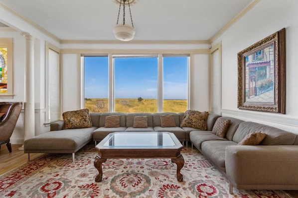 A spacious living room with a large sectional sofa, a glass-top coffee table, and a view of a grassy field through large windows. - AMAZING VIEW, 2 Homes, Hot Tubs, Steps to Beach, 8 Bedrooms, RecRoom (K&KGst) (Long Beach)