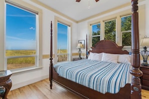 A four-poster bed with a striped bedspread, large windows with a view of a grassy field, and a ceiling fan. - AMAZING VIEW, 2 Homes, Hot Tubs, Steps to Beach, 8 Bedrooms, RecRoom (K&KGst) (Long Beach)