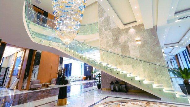 Un hall d’hôtel moderne avec un escalier en verre, un grand lustre et un coin salon doté d’une table basse.