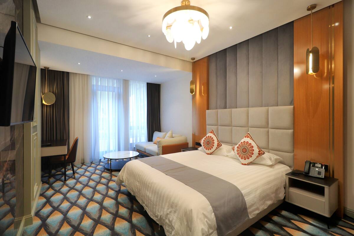 A hotel room with a large bed, a bedside table with a phone, a round table, a sofa, and a TV.