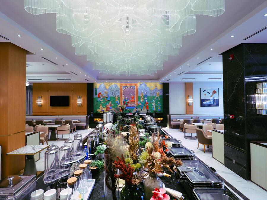 A modern hotel dining area with a long buffet table, seating arrangements, and a large chandelier.