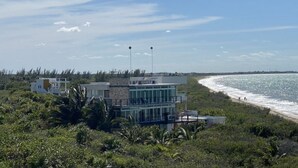 A coastal property with a modern building featuring large windows and a balcony, surrounded by lush greenery and a beach.