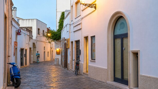 A narrow cobblestone street with buildings on either side, a scooter parked on the left, and a bicycle on the right.