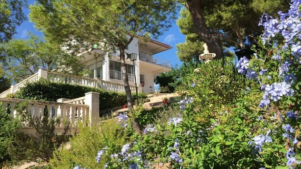 A two-story building with a balcony, surrounded by lush greenery and flowering plants.