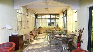 Un patio interior iluminado por el sol, con mobiliario de mimbre, una mesa de vidrio y un jarrón de flores.