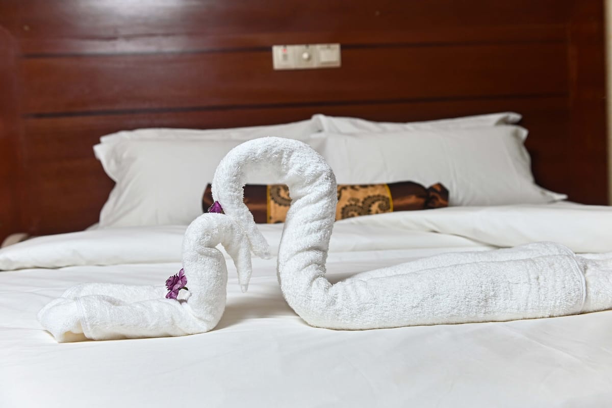 A neatly made bed with white linens and a decorative towel arrangement in the shape of a swan with a purple flower.