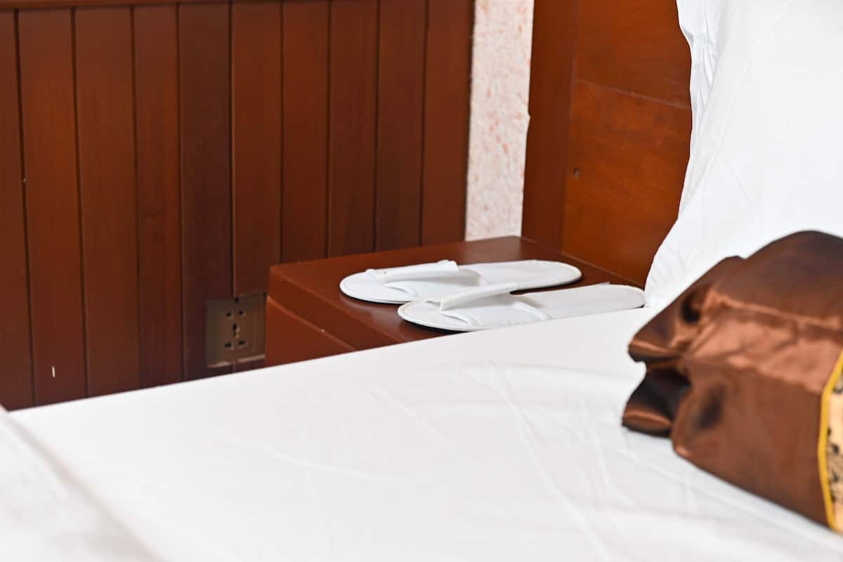 A hotel room with a wooden headboard, a bed with white bedding, a brown pillow, and a pair of white slippers on a wooden nightstand.