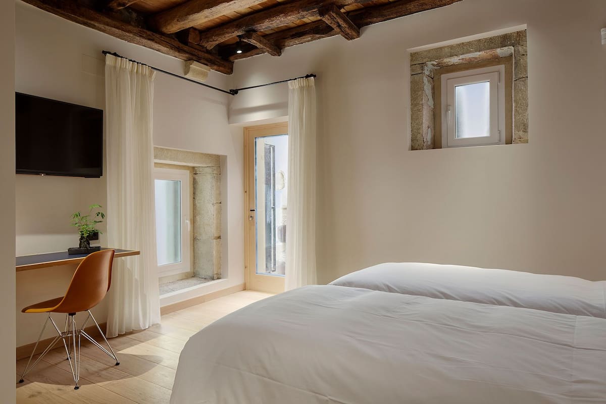 A bedroom with a bed, a desk with a chair, a TV, and a window with curtains.