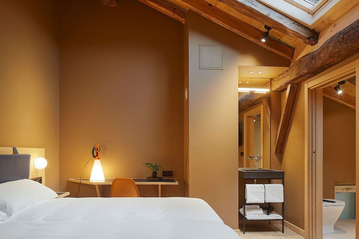 A bedroom with a skylight, a bed, a desk with a lamp, and a bathroom with a sink and towel rack.