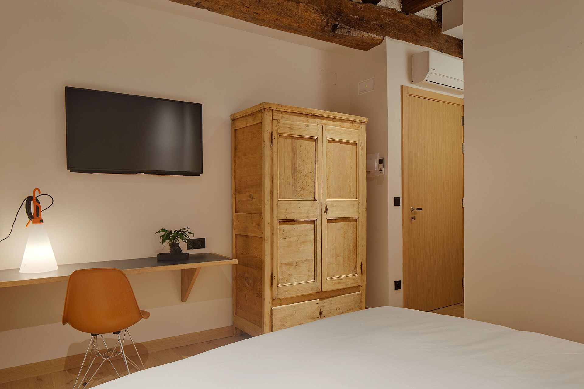 A bedroom with a bed, a desk with a chair, a television, a wardrobe, and a bedside lamp.