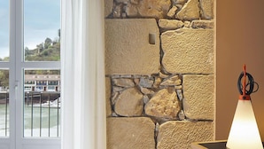 Un dormitorio con pared de piedra, una cama, una ventana con cortinas y una lámpara.