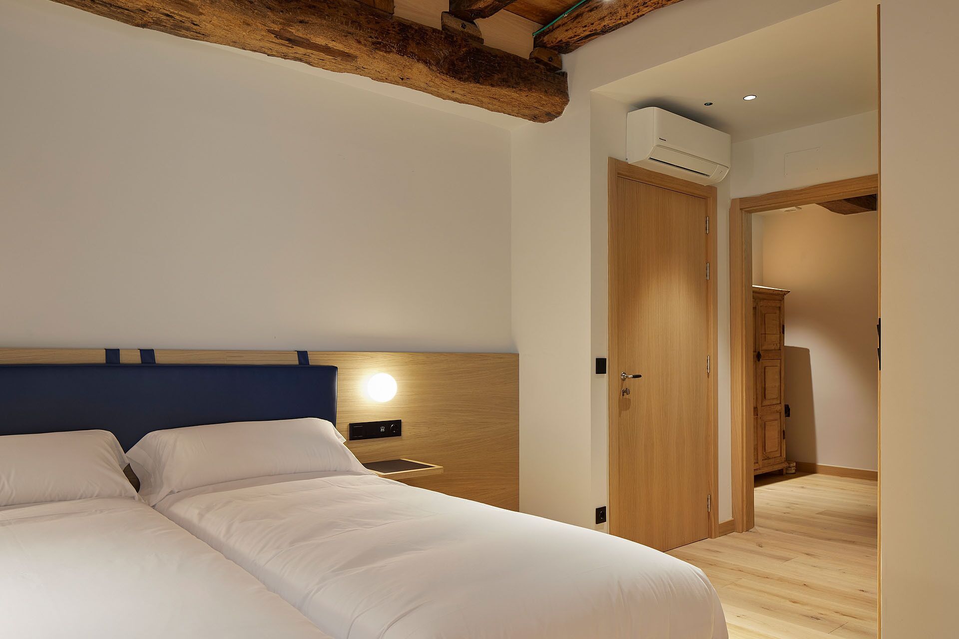 Un dormitorio con una cama grande, cabecera de madera, una mesita de noche con lámpara y una puerta que da a otra habitación.