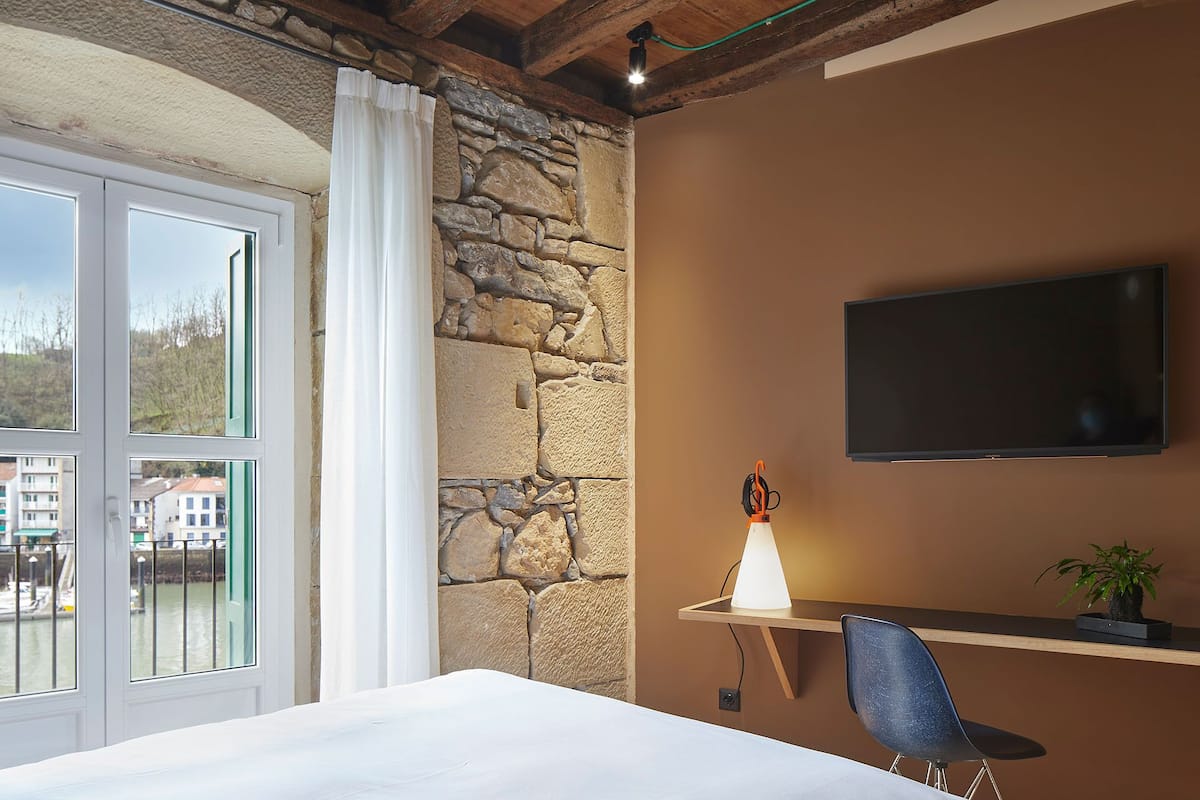 A bedroom with a stone wall, a TV mounted on the wall, a bedside table with a lamp, a chair, and a window with a view of buildings and water.
