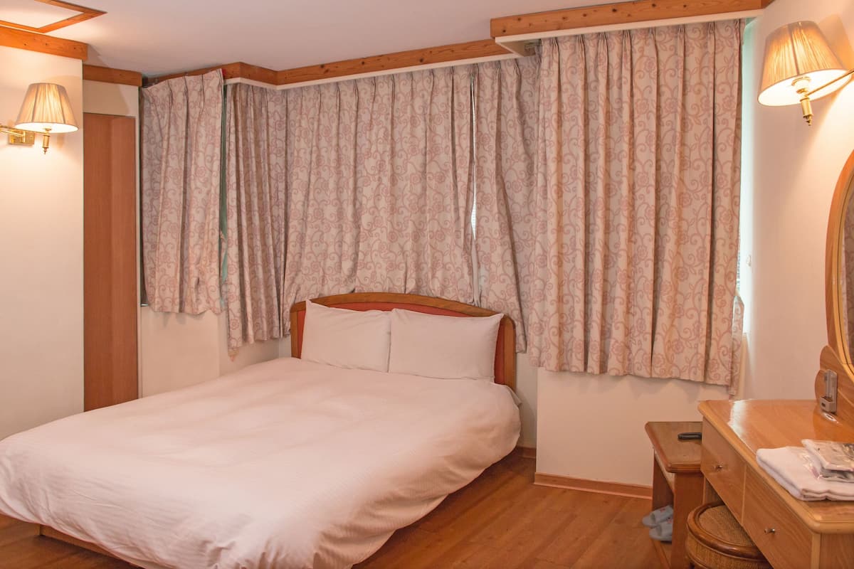 A bedroom with a wooden bed, a wooden nightstand, a mirror, and a window with patterned curtains.