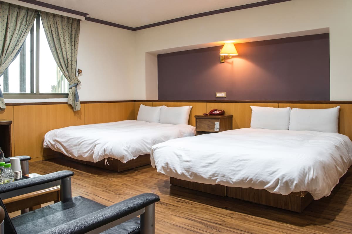 A hotel room with two beds, a desk, and a chair. There is a window with curtains, a wall-mounted lamp, and a bedside table with a phone.