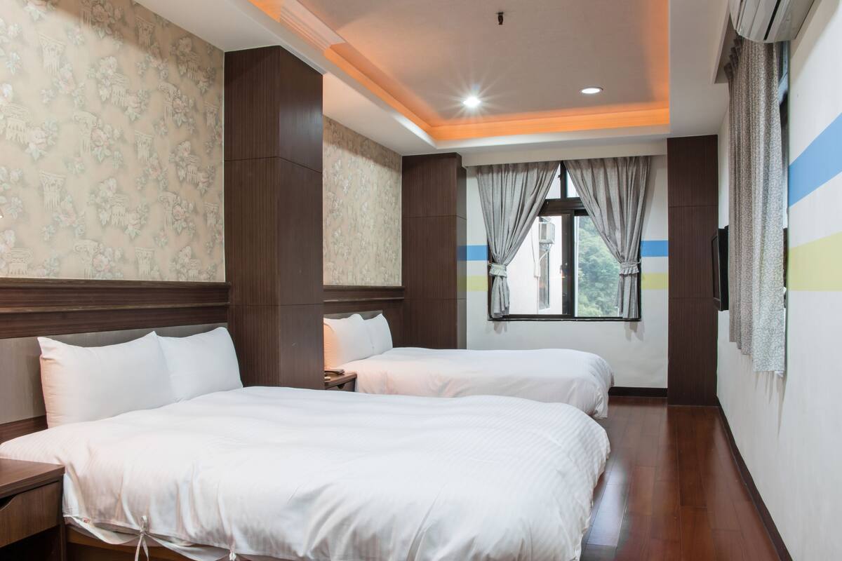 A hotel room with two beds, wooden flooring, a large window with curtains, and a TV mounted on the wall.