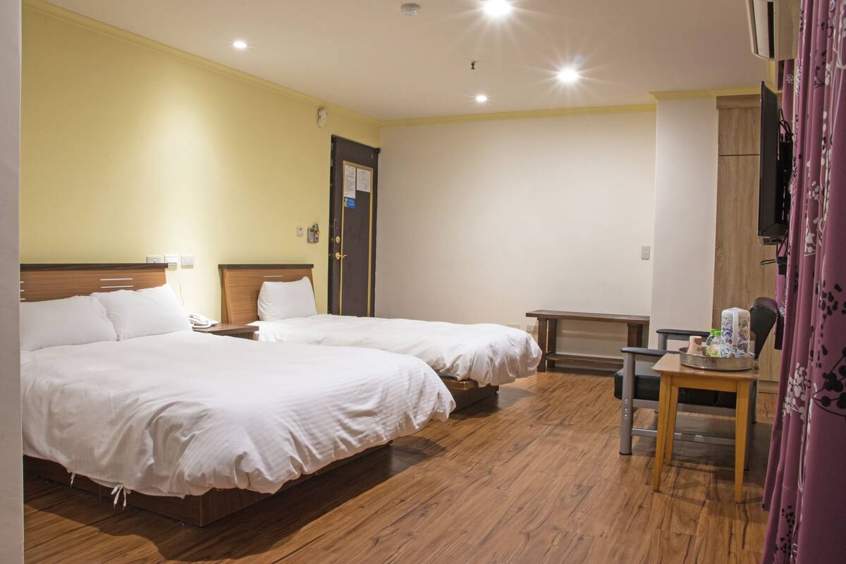 A hotel room with two beds, wooden flooring, a small table with a basket, and a television mounted on the wall.