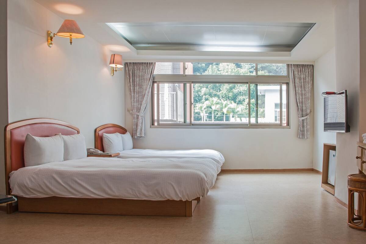 A hotel room with two beds, a skylight, a large window with curtains, and a wall-mounted TV.