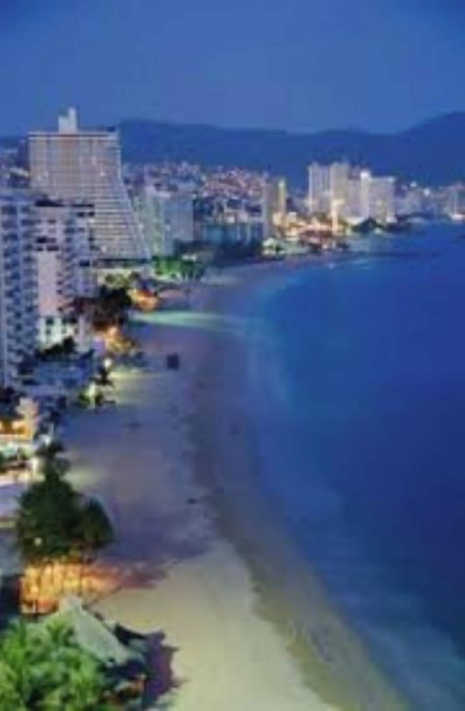 A coastal cityscape at night with high-rise buildings, a beach, and a clear sky.
