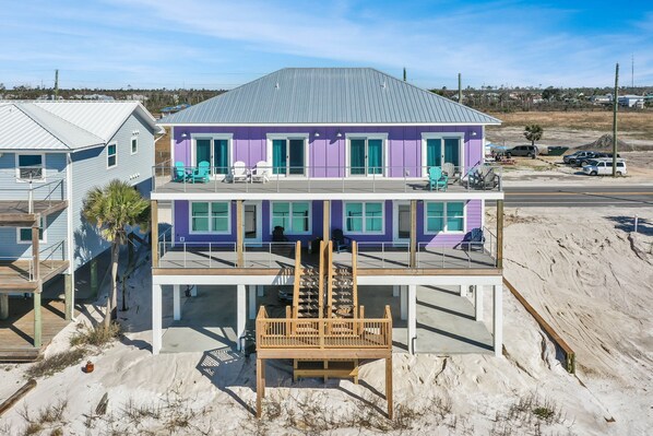 Una casa de playa de dos pisos con un balcón, una terraza de madera y una zona de arena.