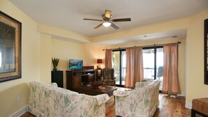 A living room with floral-patterned sofas, a wooden coffee table, a television, and a ceiling fan. - Beach View, Next to Pool, 2nd floor- Club at Mexico Beach 2J (Mexico Beach)