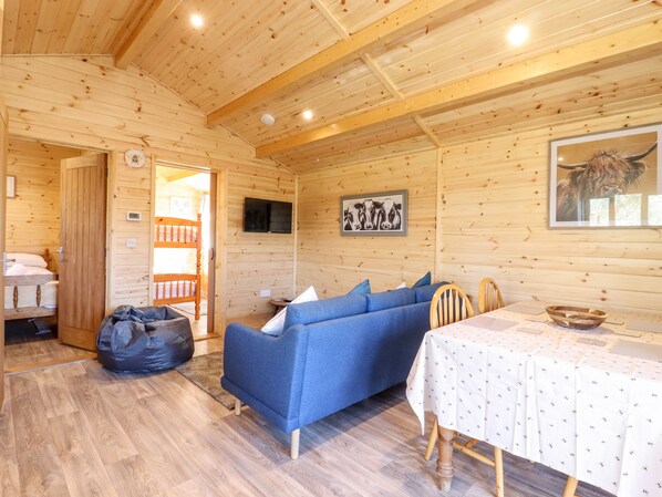 A cozy cabin interior with a blue sofa, a dining table with a white tablecloth, and a wooden ceiling. - Clover Lodge (Leiston)