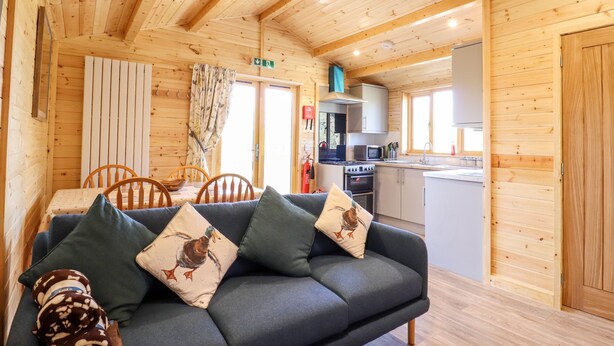 A cozy cabin interior with a sofa, dining table, and kitchen area. - Clover Lodge (Leiston)