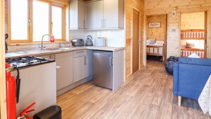 A wooden cabin with a kitchen featuring a stove, sink, and cabinets. There is a blue couch and a bed with a wooden headboard in the living area. - Clover Lodge (Leiston)