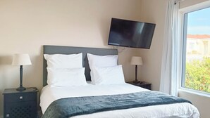 A hotel room with a bed, two bedside tables with lamps, a flat-screen TV mounted on the wall, and a window with a view of buildings and greenery. - Vitamin Sea (Plettenberg Bay)