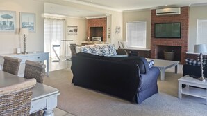 A living room with a sofa, a dining table, wicker chairs, a fireplace, and a TV mounted on the wall. - Vitamin Sea (Plettenberg Bay)