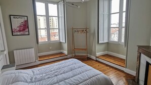 A bedroom with a bed, a radiator, a wooden floor, a window with a view, and a painting on the wall. - Beautiful renovated apartment, 5 min from the beach! (BIARRITZ)