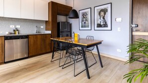 Une cuisine moderne avec des meubles en bois, un lave-vaisselle en acier inoxydable, un réfrigérateur noir et une table à manger avec des chaises noires. Deux œuvres d’art encadrées ornent le mur, et une suspension lumineuse est accrochée au-dessus de la table.