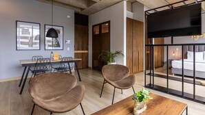 Un intérieur moderne avec une table à manger en bois, deux chaises marron, une table basse agrémentée d’une plante, et un lit avec un téléviseur en arrière-plan.
