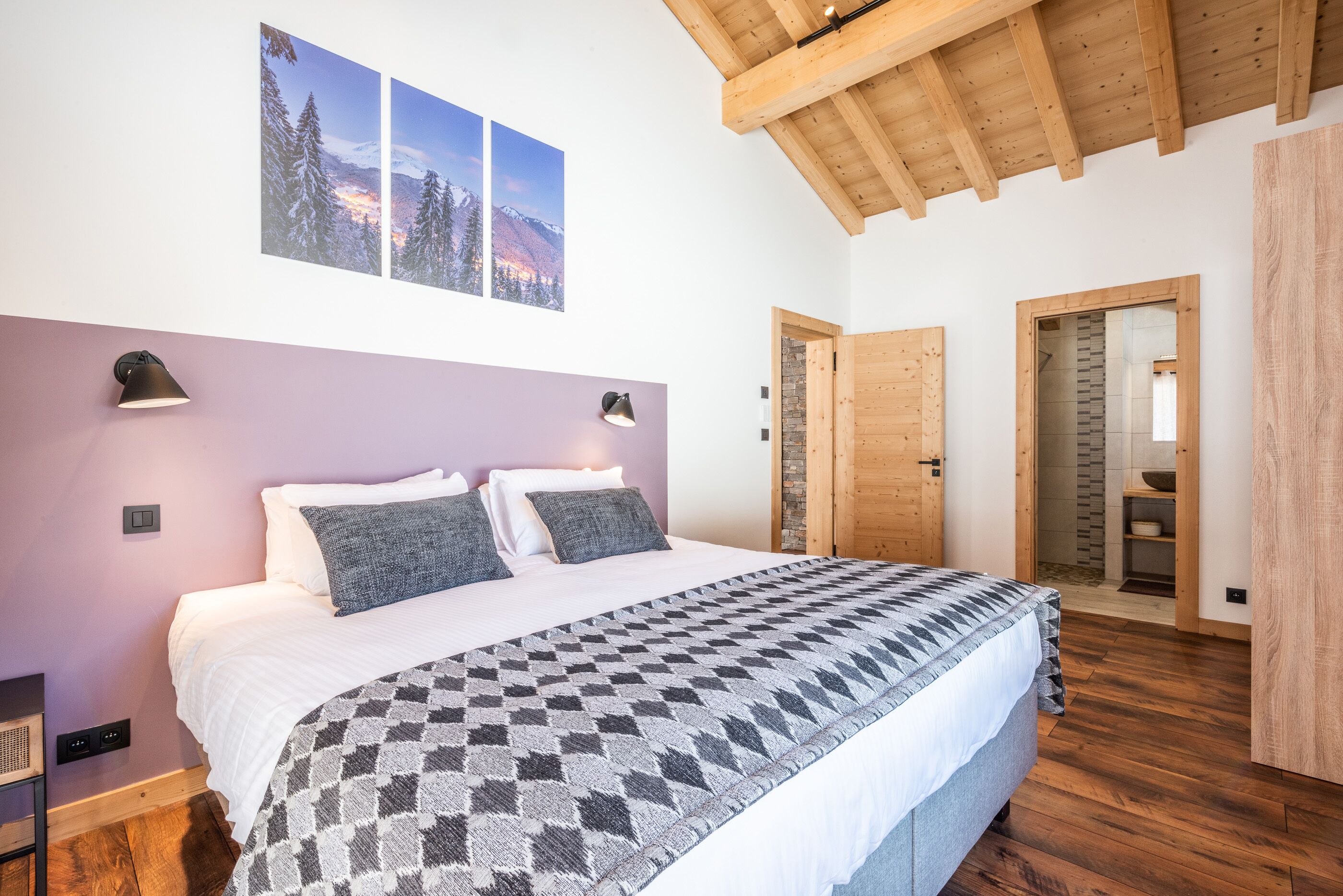 A bedroom with a large bed, two wall-mounted lamps, a wooden ceiling, and a view of a snowy mountain landscape through the window.