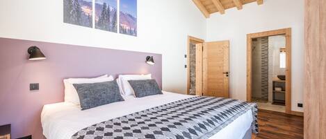 A bedroom with a large bed, two wall-mounted lamps, a wooden ceiling, and a view of a snowy mountain landscape through the window.