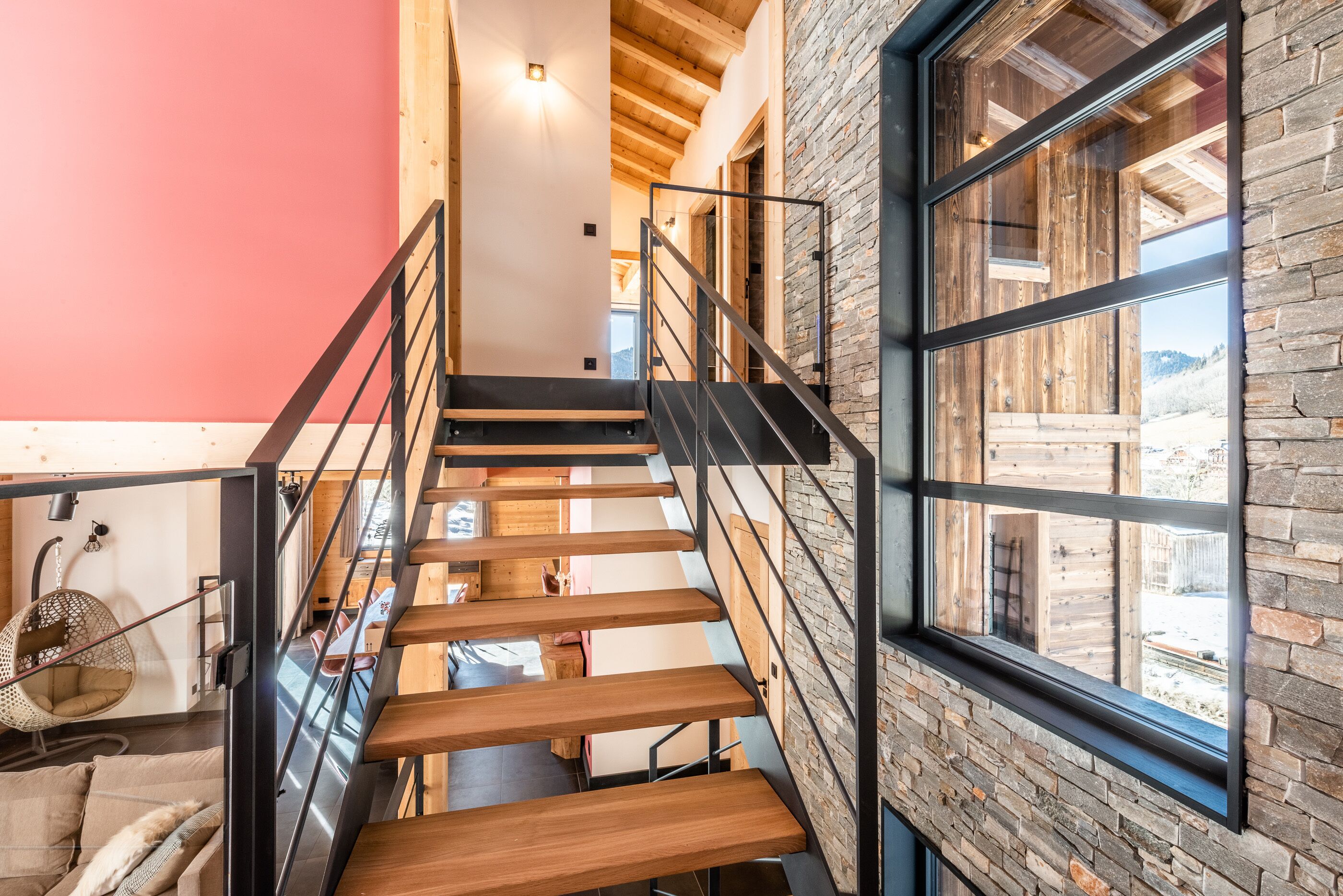 A wooden staircase with black railings, a hanging chair, and a view of the outdoors through a large window.