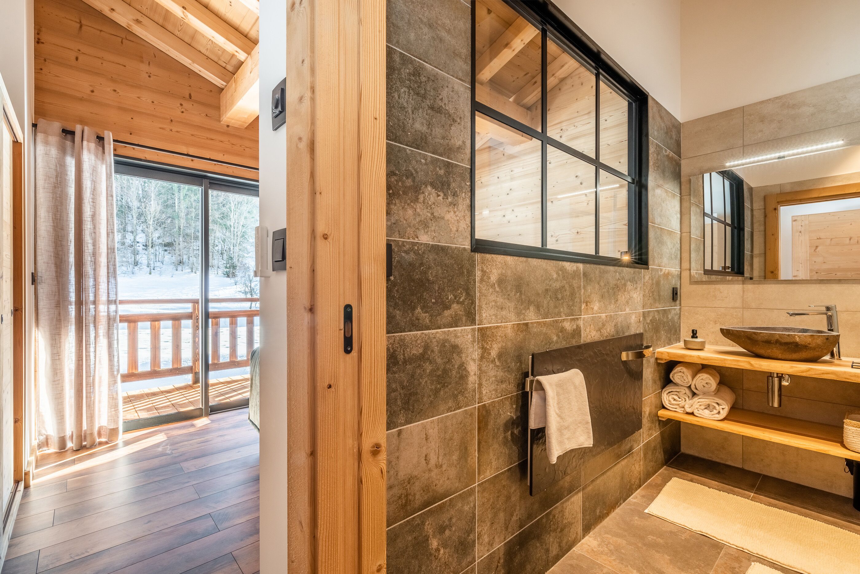 Ein modernes Badezimmer mit Holzboden, Steinfliesen, einem großen Fenster und einem hölzernen Waschtisch mit Waschbecken und Handtuchhalter.