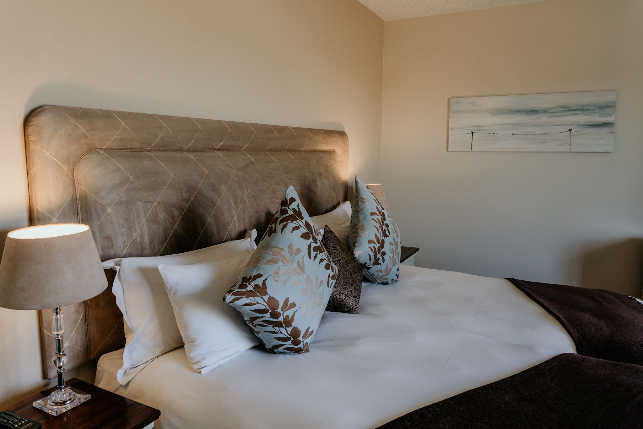 A neatly made bed with a headboard, a bedside lamp, and a painting on the wall.