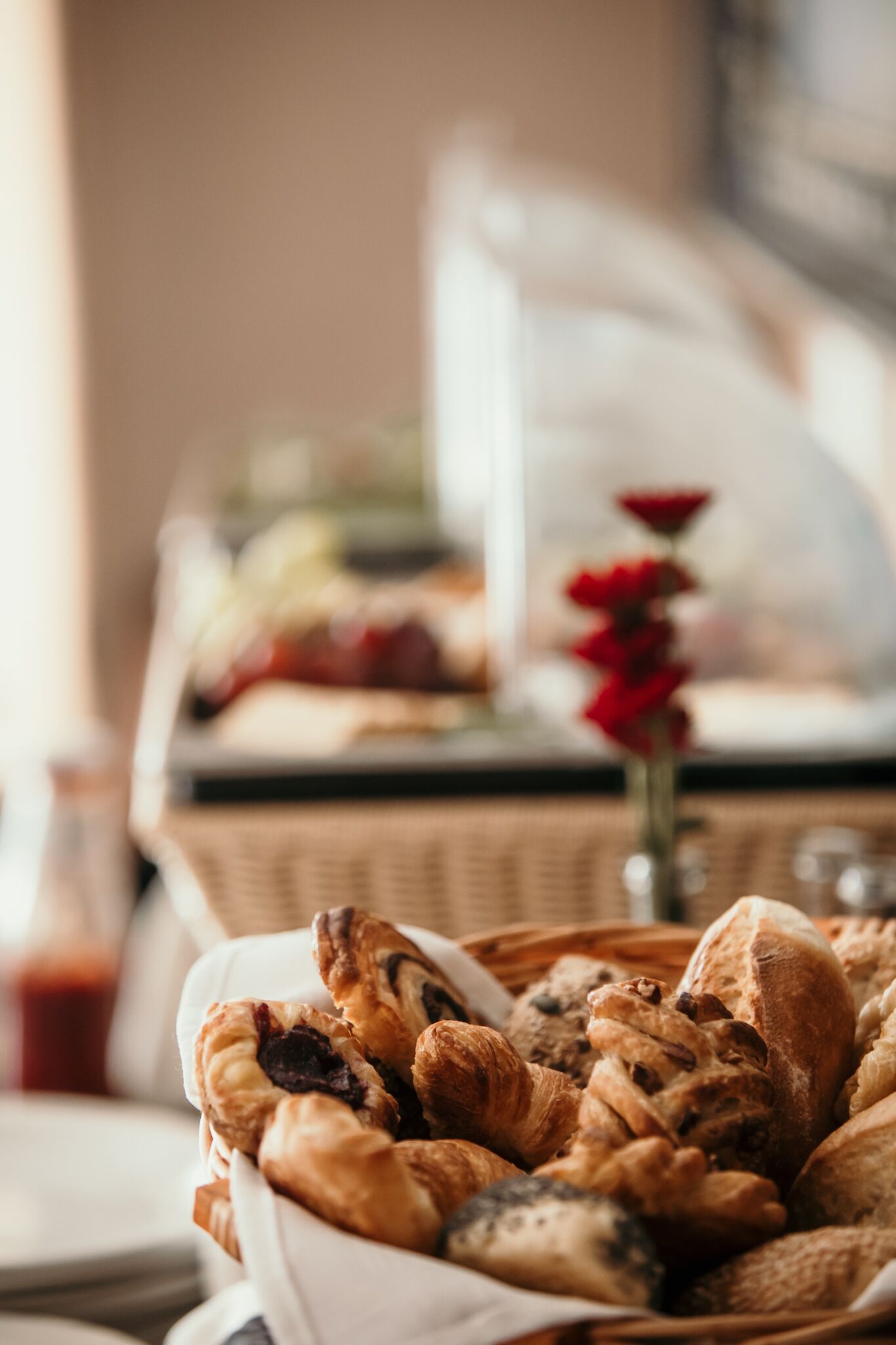 A basket of assorted pastries and bread, a glass of red beverage, and a vase with red flowers on a table.