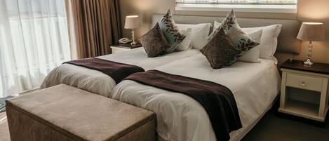 A double bed with a dark throw blanket, two bedside tables with lamps, a nightstand, a window with blinds, and a view of buildings outside.