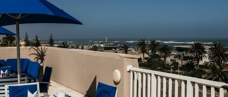 Eine Terrasse mit Esstisch, einem blauen Sonnenschirm und Blick auf das Meer.