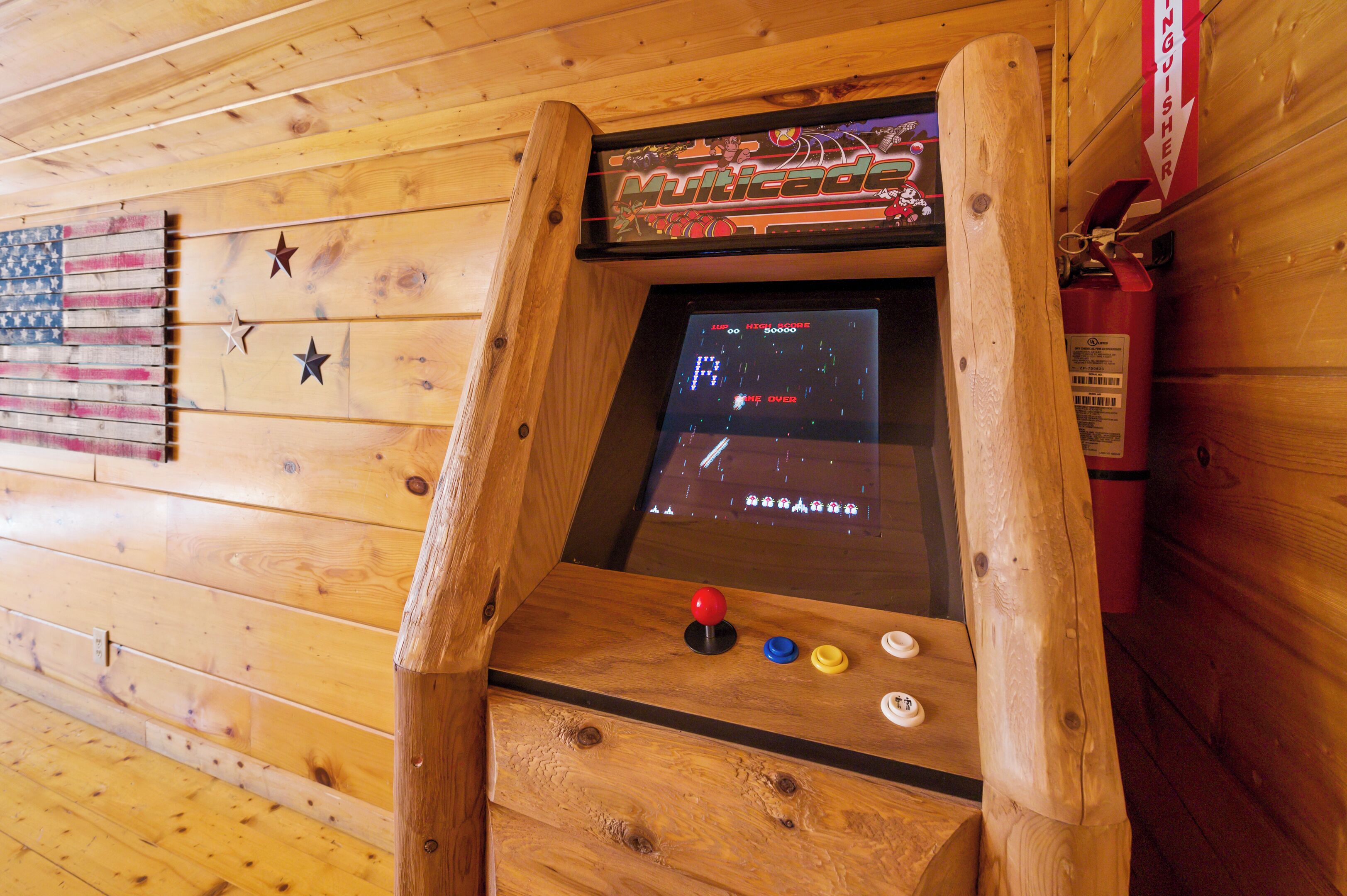 Une borne d’arcade en bois dotée d’un écran affichant un jeu de course, le tout installé devant un mur en bois orné d’un drapeau américain et d’étoiles.