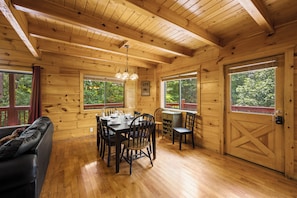 Interior de uma cabana de madeira com uma mesa de jantar posta para quatro, rodeada por cadeiras, e um sofá em primeiro plano.
