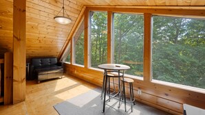 En inredning med träpanel, en sovplats på ett loft, en soffa och ett matbord med stolar. Stora fönster ger utsikt över skogen.