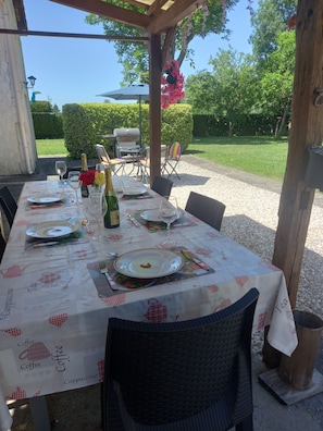 Un espace de restauration en plein air avec une table dressée pour deux personnes, agrémentée d’une nappe ornée de motifs de café et de thé, d’une bouteille de champagne et d’un barbecue en arrière-plan.