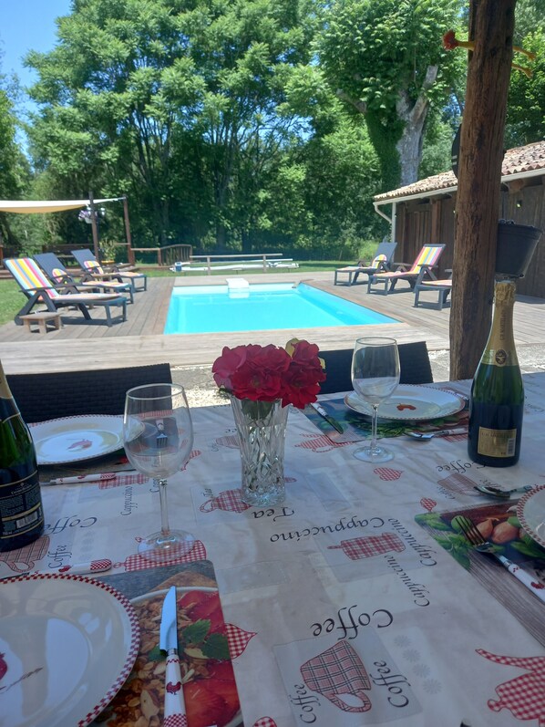 Un espace repas aménagé au bord de la piscine, avec une table dressée pour deux personnes, agrémentée d’une bouteille de champagne, d’un vase de roses rouges et d’un menu sur le thème du café.