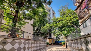 Une cour avec un sol à carreaux, entourée de bâtiments dotés de balcons et d’un arbre qui offre de l’ombre.